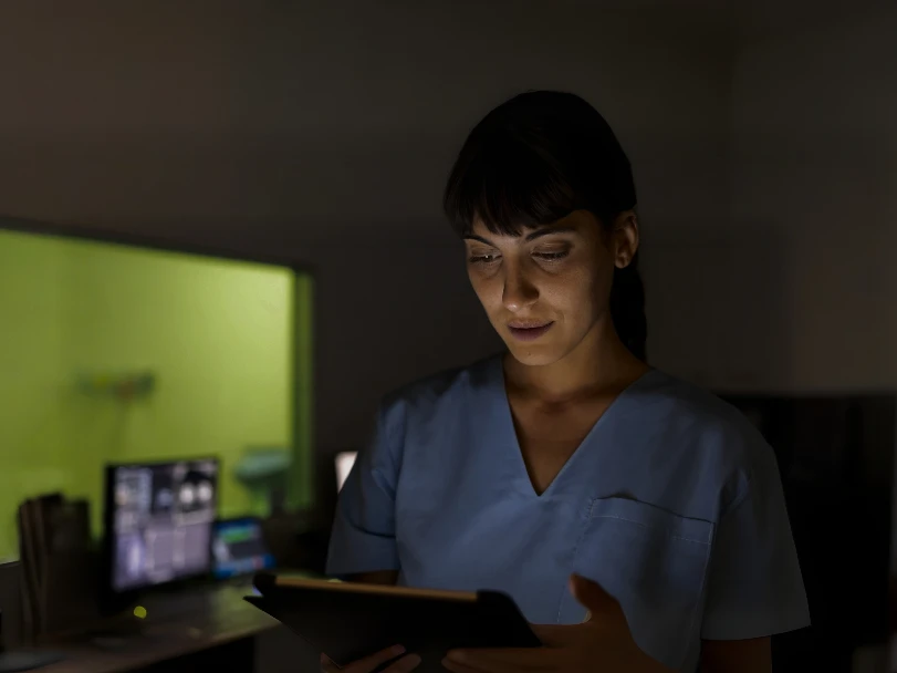 A nurse reading a tablet at night, a patient monitor lit behind her.