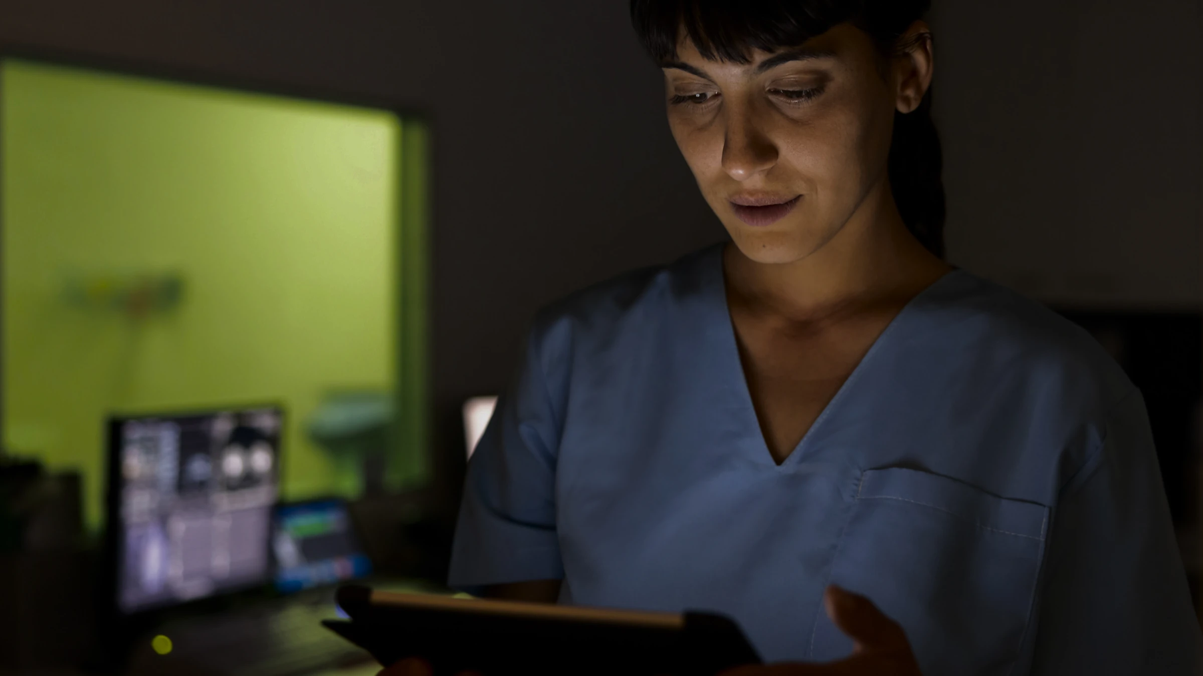 A nurse reading a tablet at night, a patient monitor lit behind her.